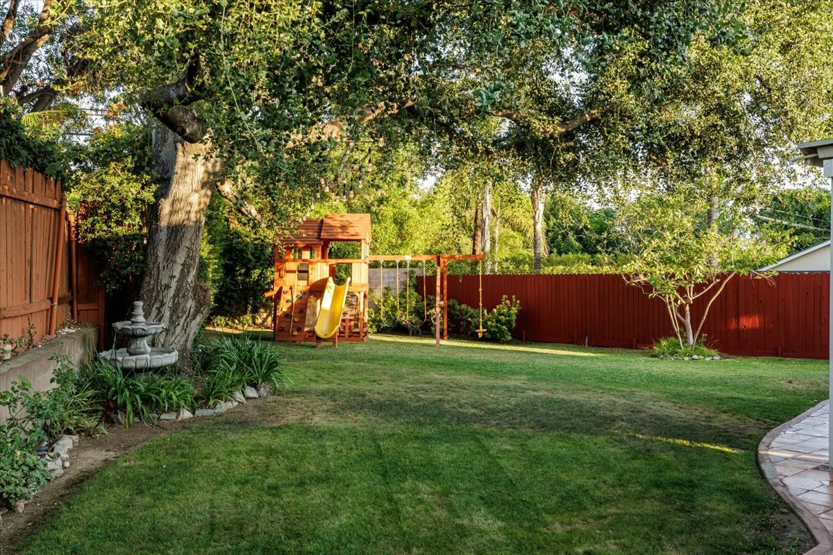 Backyard with large tree providing shade, lawn, wooden playset with slide and swings, flower bed with plants and a stone fountain, red wooden fence, and a curved stone pathway.