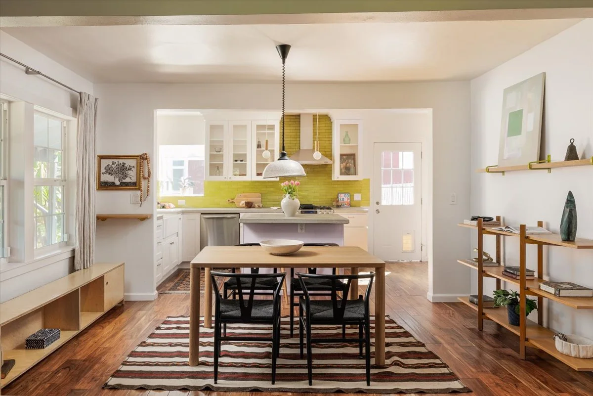 Open-concept kitchen and dining area with white walls, wooden flooring, a dining table with four chairs, and a kitchen with white cabinets, a yellow backsplash, and a central island. Decor includes a vase of flowers on the table, framed artwork, and 