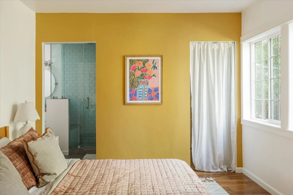 A bedroom with a bed, beige quilt, and pillows, a yellow accent wall with a framed floral painting, a white window with curtains, and an open doorway leading to a bathroom with a glass shower door.