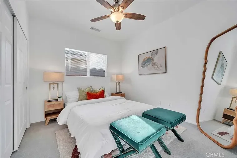 A bright white bedroom with a bed, two nightstands with lamps, artistic wall decor, a ceiling fan, and a full-length mirror.