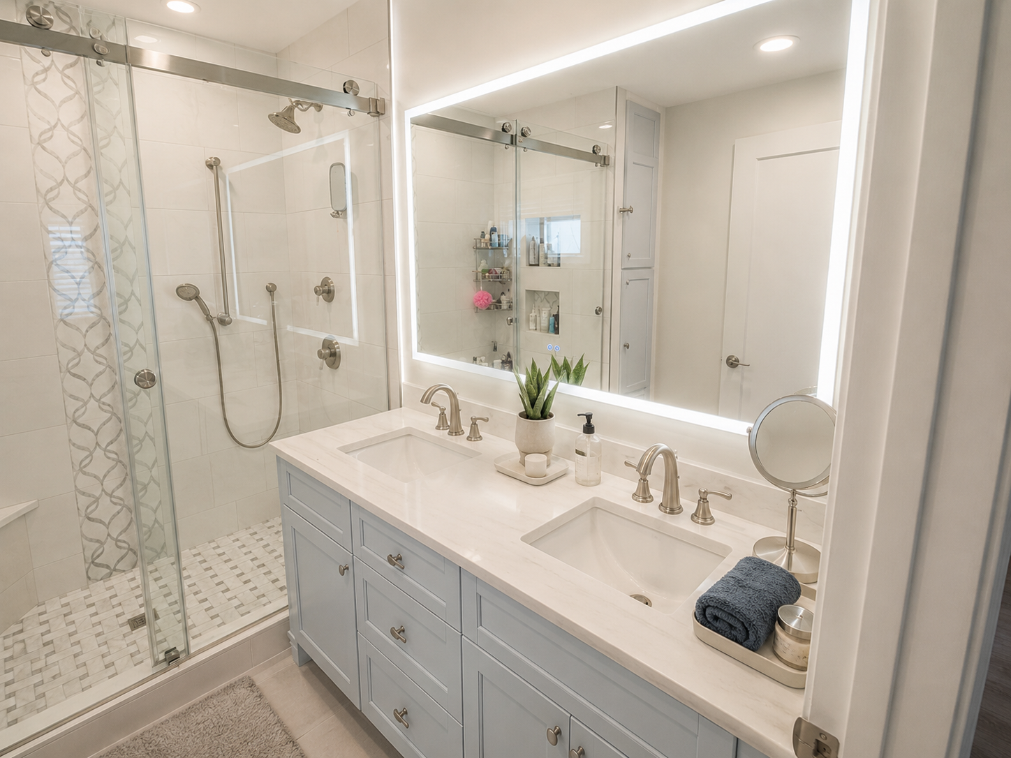 Modern bathroom with a double vanity with a light blue cabinet, white marble countertop, and two sinks. There is a large backlit mirror, a potted plant, and bathroom essentials on the counter. The shower has a glass door, and white tiles with a decor