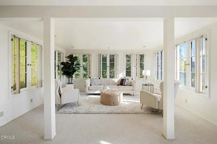 Bright living room with white walls, large windows, and white furniture including sofas and armchairs, with a round ottoman, indoor plant, and a side table with a lamp.