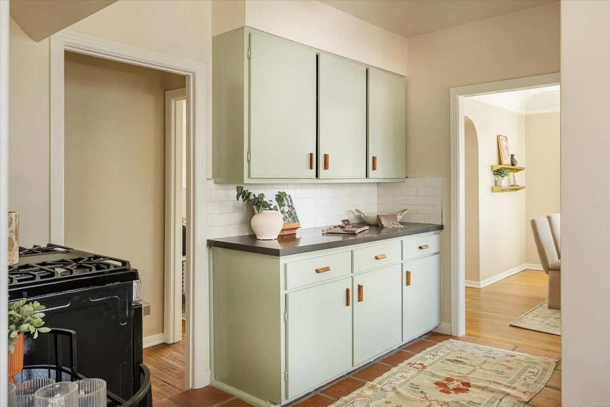 A kitchen corner with pastel green cabinets, a dark countertop, and white subway tile backsplash. There is a potted plant, a small framed picture, and some decorative items on the counter. Adjacent to the kitchen is a room with a beige chair and wall