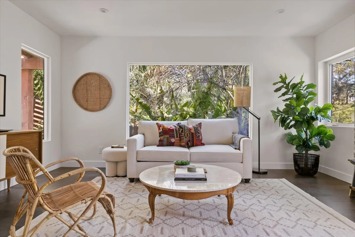 Bright living room with white walls, large windows showing greenery outside, a white sofa with colorful pillows, a round marble-top coffee table, wicker chairs, a tall floor lamp, and a potted plant.