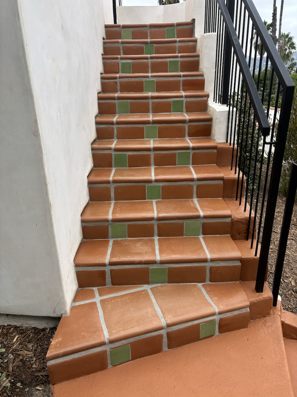 Outdoor staircase with brown and green tiles, black metal railing on right side, white walls on the left, and orange concrete at the bottom.