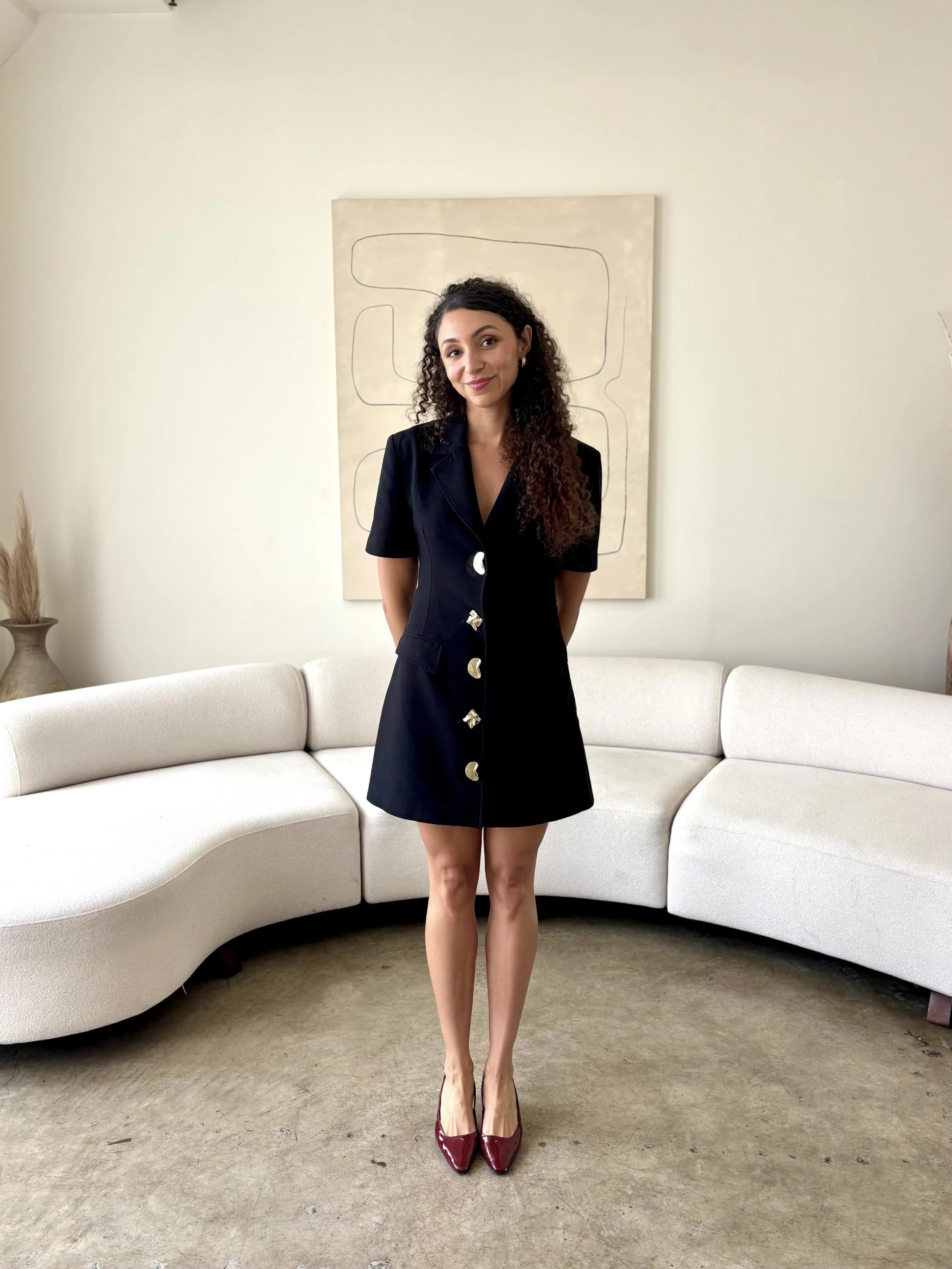 A woman with curly brown hair, wearing a navy blue dress with large metallic buttons, stands in front of a cream-colored sectional sofa, with a beige abstract artwork on the wall behind her and a vase with dried plants on the left.