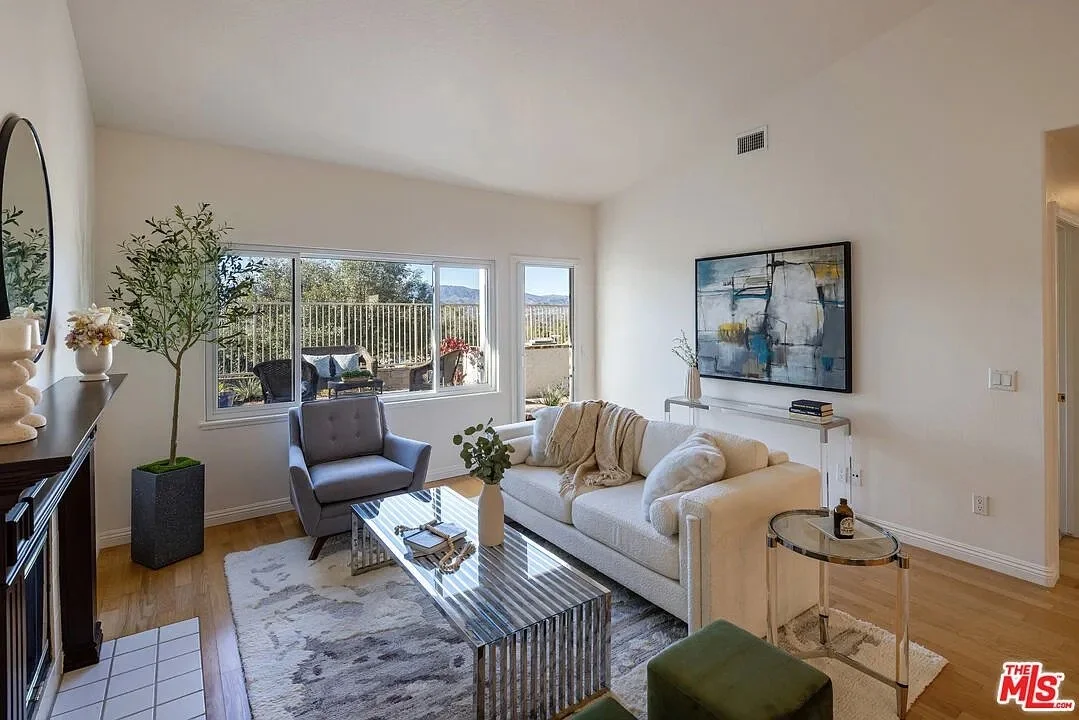 Bright living room with white sofa, gray armchair, glass-top coffee table, green ottomans, and a large window overlooking a balcony or outdoor space.