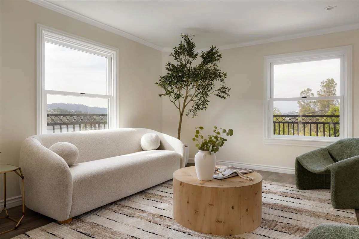 A bright, minimalist living room with two large windows, a white sofa with round pillows, a green armchair, a round wooden coffee table with a white vase and greenery, a tall potted tree, and a beige striped rug.