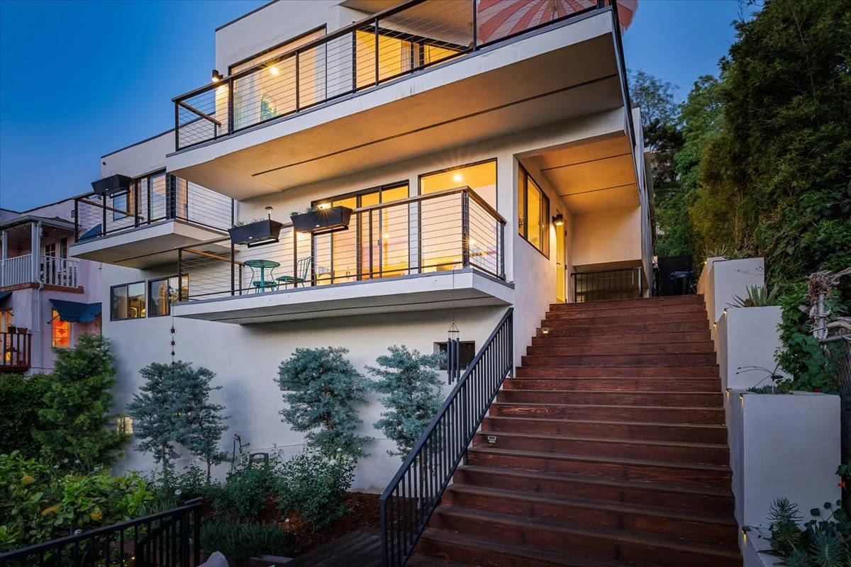 Modern multi-story house with large glass windows and balconies, wooden stairs leading up to the entrance, surrounded by greenery and outdoor lighting, during twilight.