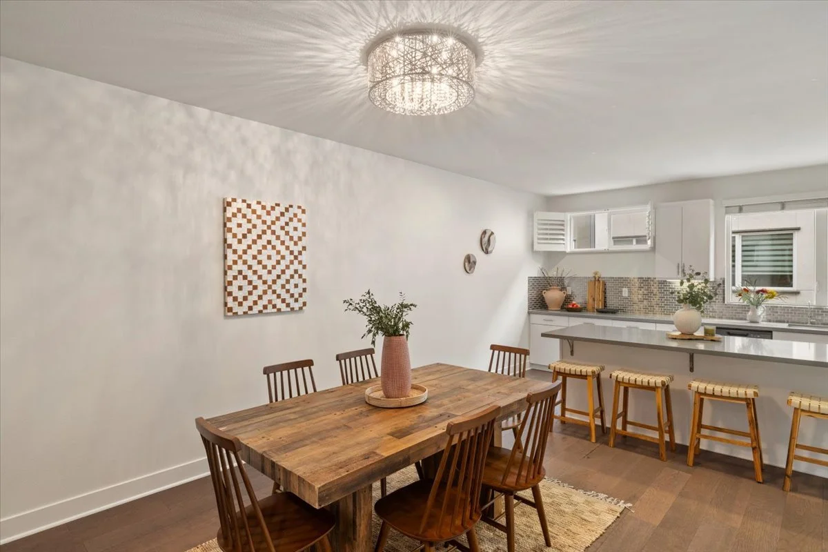 Open-concept dining and kitchen area with a rustic wooden dining table, six matching chairs, a modern chandelier, and a white kitchen counter with bar stools, decorated with vases and plants, and a wall art piece.