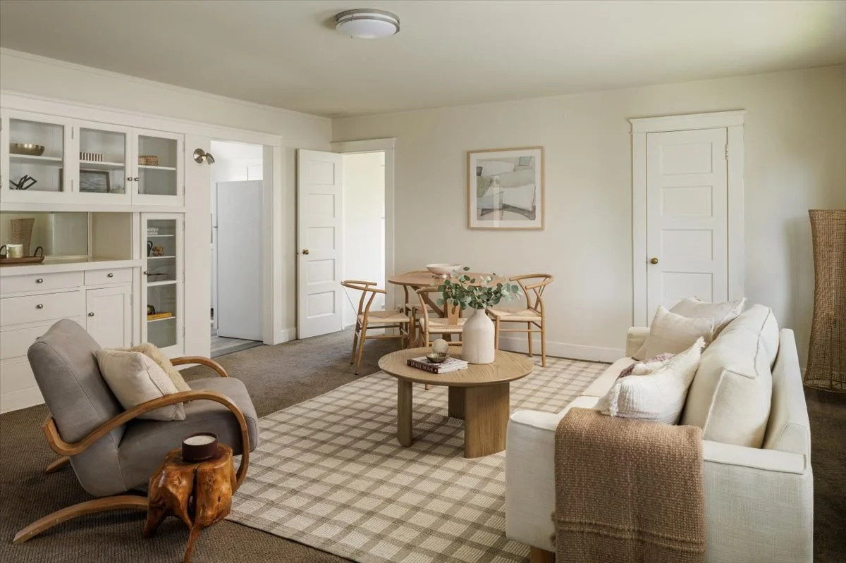 Living room with white couch, gray armchair, wooden coffee table with vase and books, dining table with four chairs, built-in white cabinet, and a doorway on the left. Decor includes a framed picture and a woven room divider.