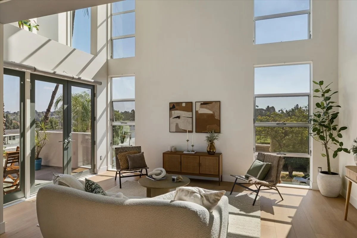 Bright living room with large windows and glass door leading to a balcony, modern furniture, abstract wall art, potted plants, and natural sunlight.