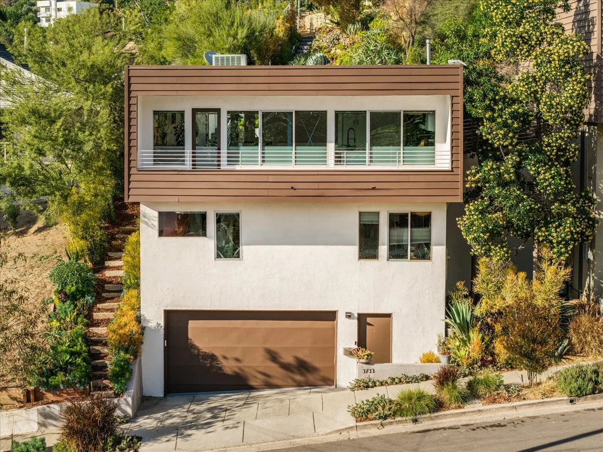 A modern three-story house with a brown garage door and second-floor balcony, surrounded by lush greenery and plants.