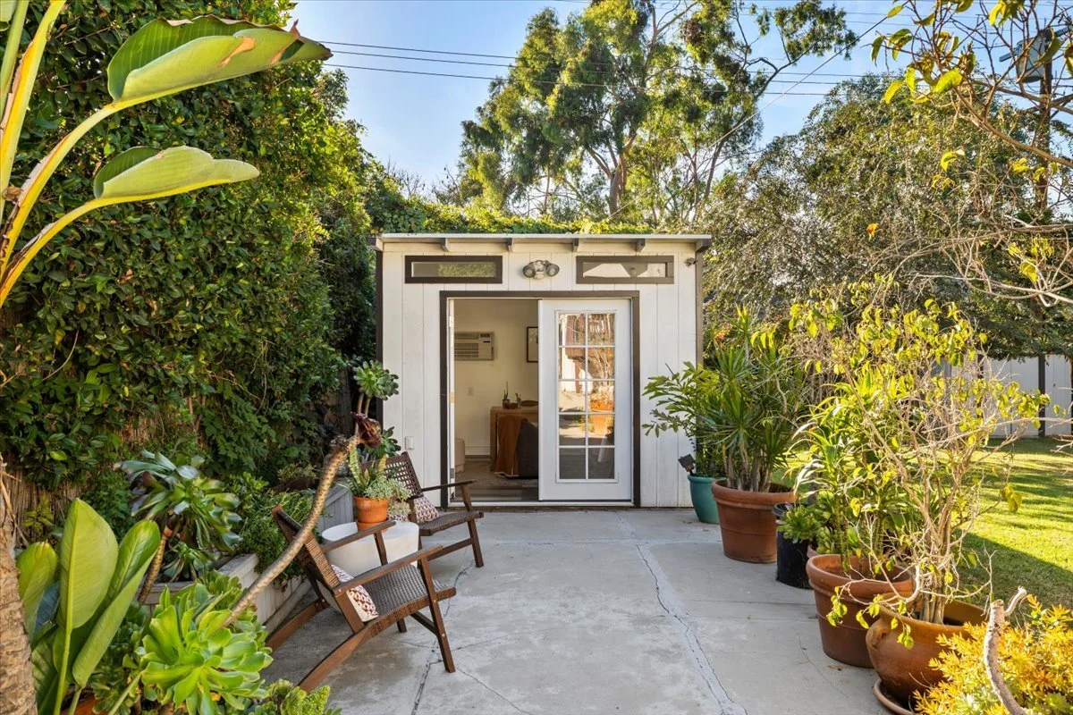 Small backyard patio area with concrete surface, surrounded by potted plants and bushes, with a white shed featuring a glass-paneled door open leading into the house. There are two wooden benches with cushions on the left side of the patio.