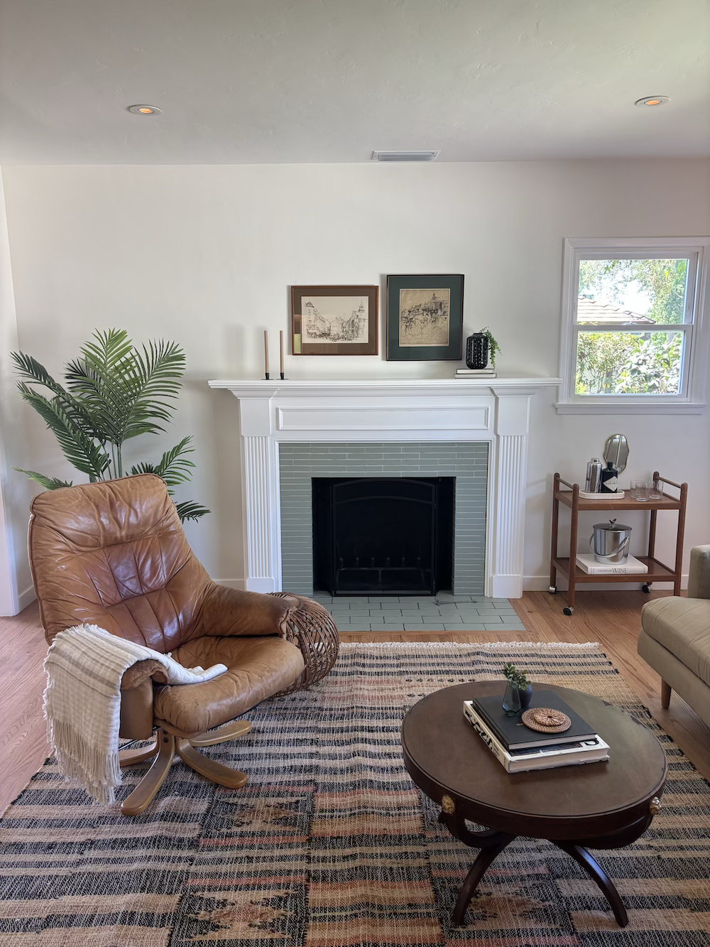 Living room with a leather armchair, a fireplace with framed art above, a window, a wooden side cart with wine accessories, a striped area rug, and a coffee table with books and decor.