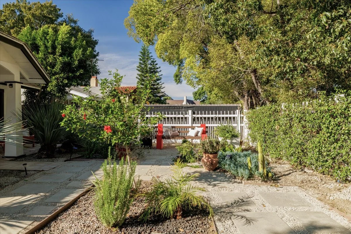 A backyard garden with a paved pathway, green bushes, and trees. There is a white fence in the background with outdoor seating and decorative pillows. Potted plants and flowering plants are also visible.