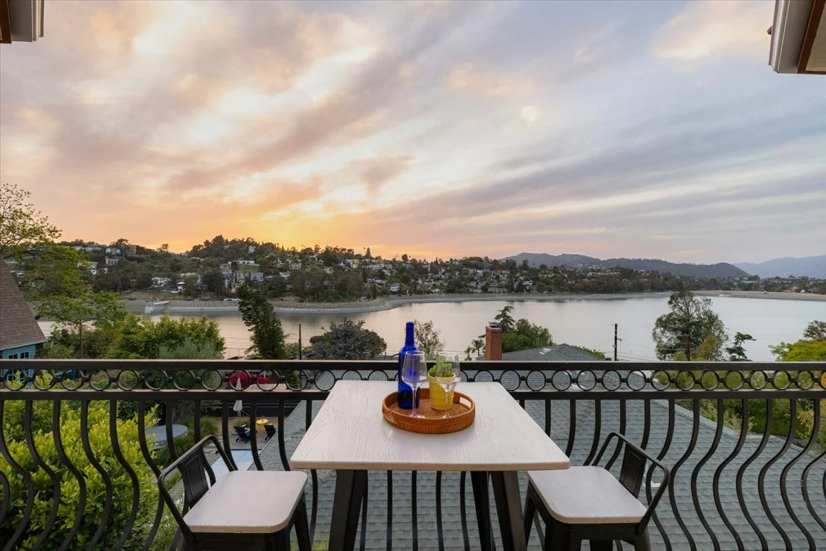 View from a balcony overlooking a river with houses and trees on the opposite bank during sunset. There is a table with a blue bottle, a glass of lemonade, and a potted plant on it, with two chairs nearby.