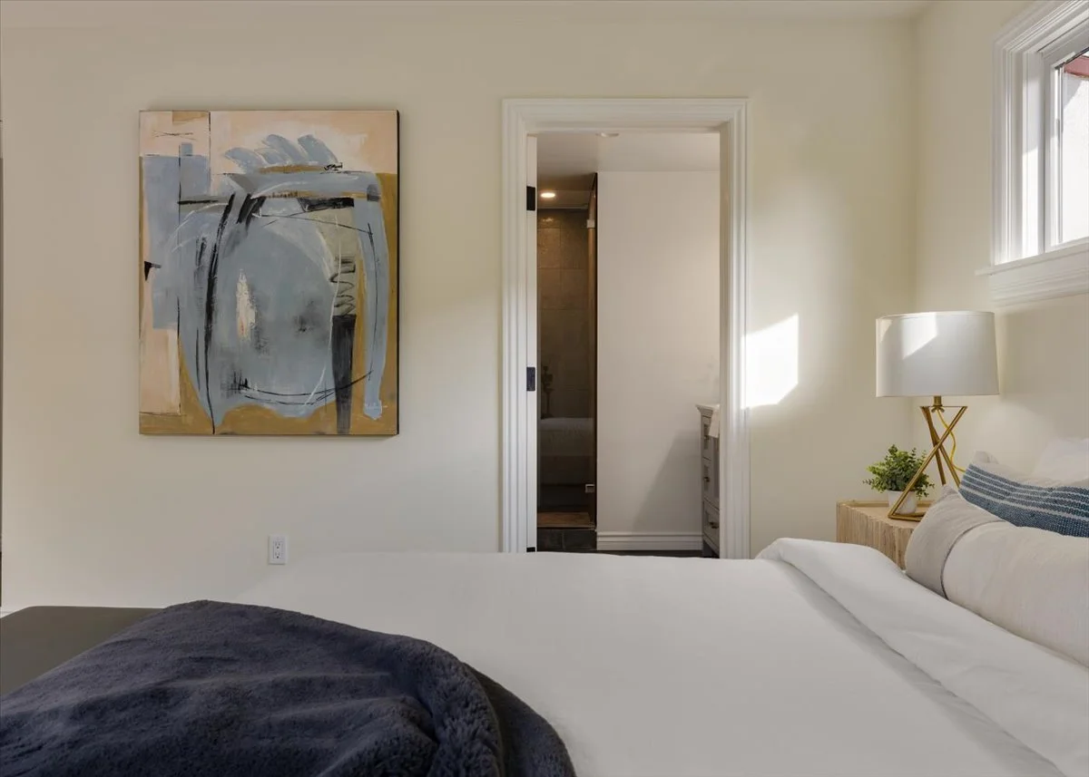 A bedroom with a bed, a window, a lamp on a bedside table, and an abstract painting on the wall, with a doorway leading to a bathroom.