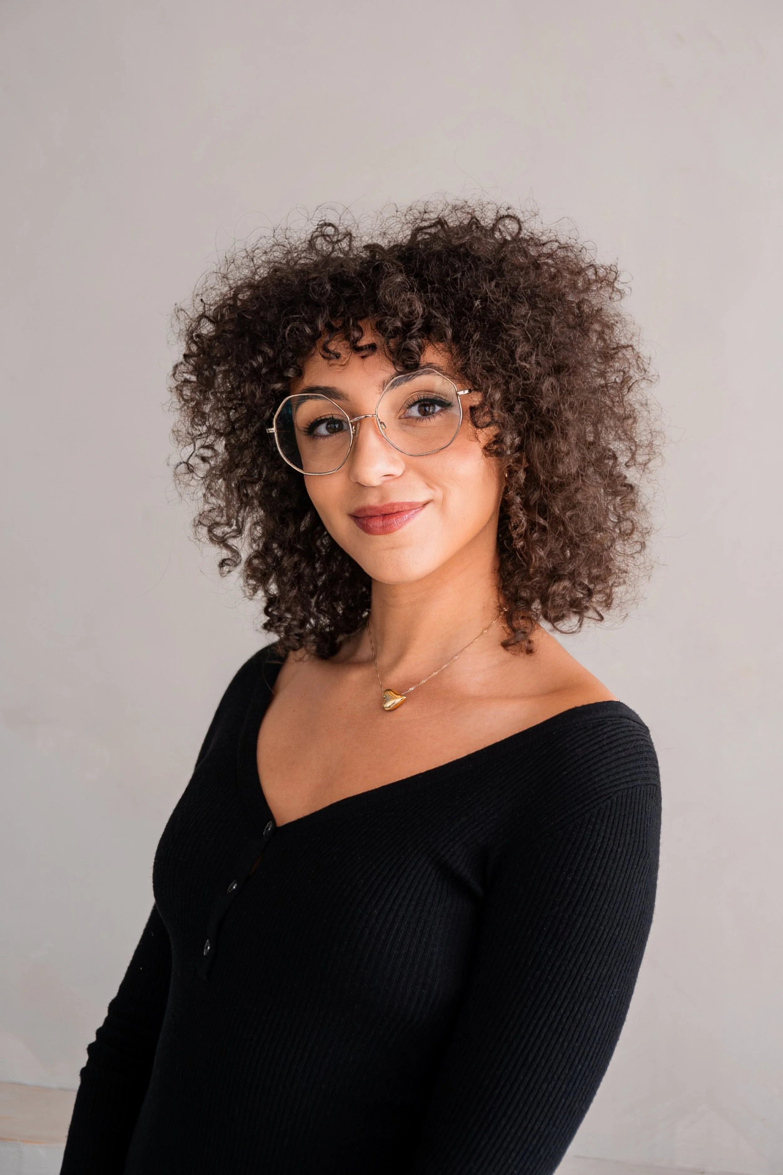 A woman with curly hair wearing glasses and a black top, smiling, against a plain background.