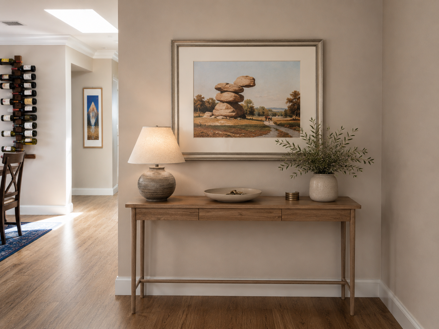 Interior scene with a wooden console table against a light-colored wall, decorated with a table lamp, a ceramic vase with greenery, a shallow bowl, and a small stack of coins.Above the table, a large framed painting of a rock formation in a landscape
