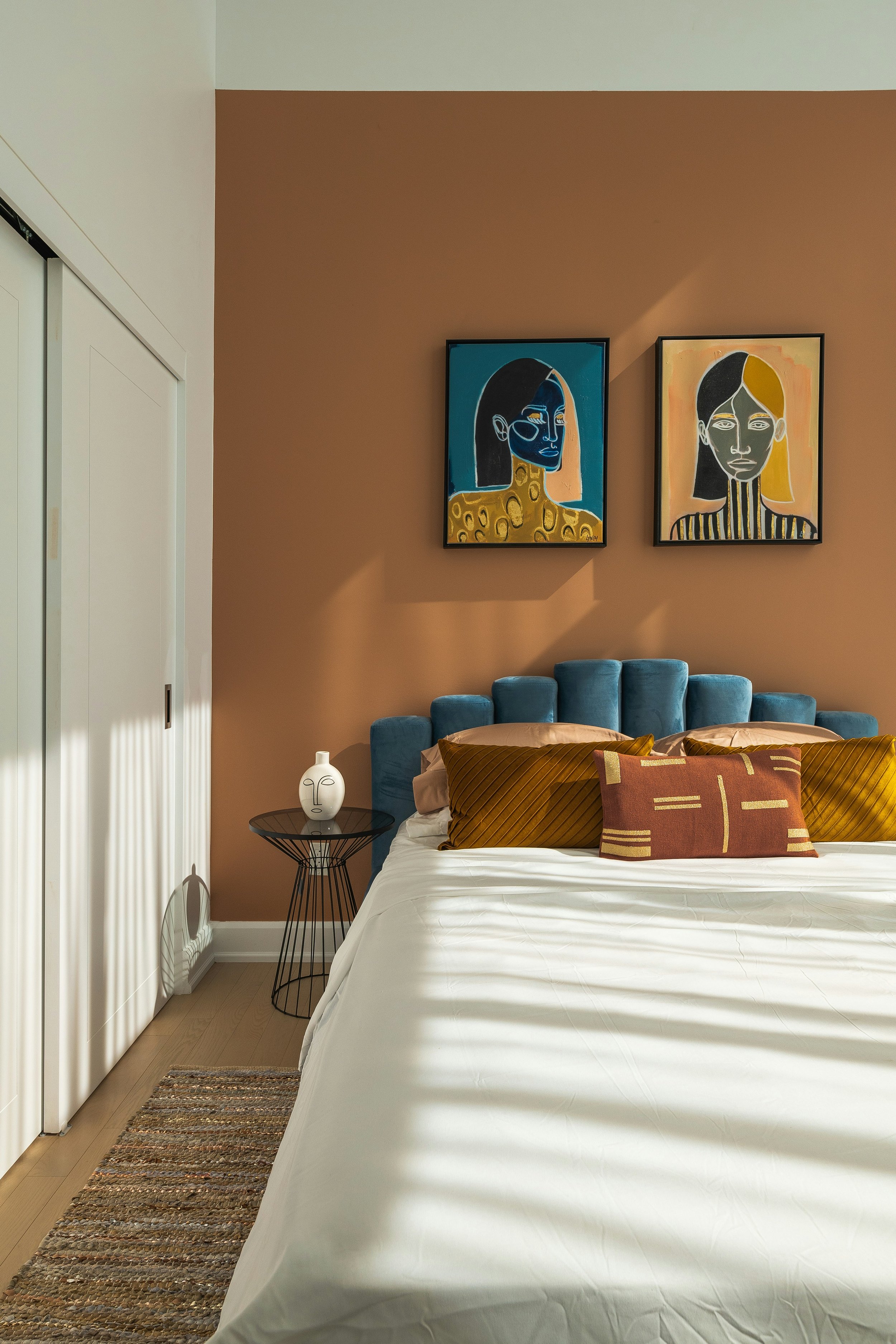 A bedroom with tan and white walls, featuring two abstract portrait paintings on the wall above a plush blue headboard. The bed has white sheets and decorative pillows in brown and yellow tones. A small black wire side table next to the bed holds a white ceramic vase with a face design. A multicolored woven rug lies on the wooden floor at the foot of the bed, and a white closet with sliding doors is partially visible on the left.