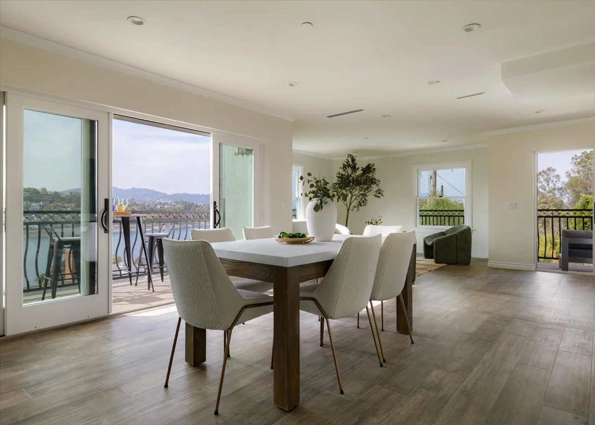 Empty dining room with large window and sliding glass door leading to balcony, view of distant hills, white table with plant centerpiece, and adjacent living area with windows and chairs.