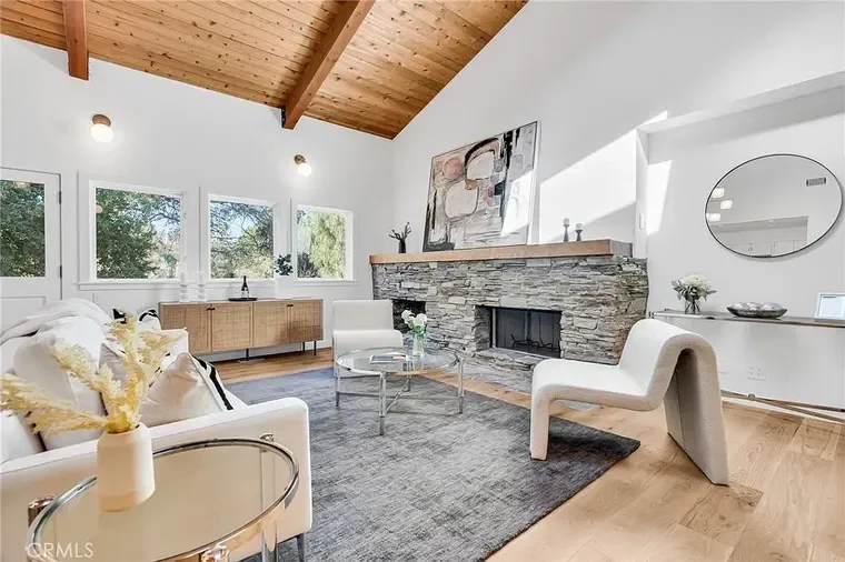Living room with white sofa, white armchair, modern fireplace with stone facade, large windows, wooden ceiling, and minimal decor.