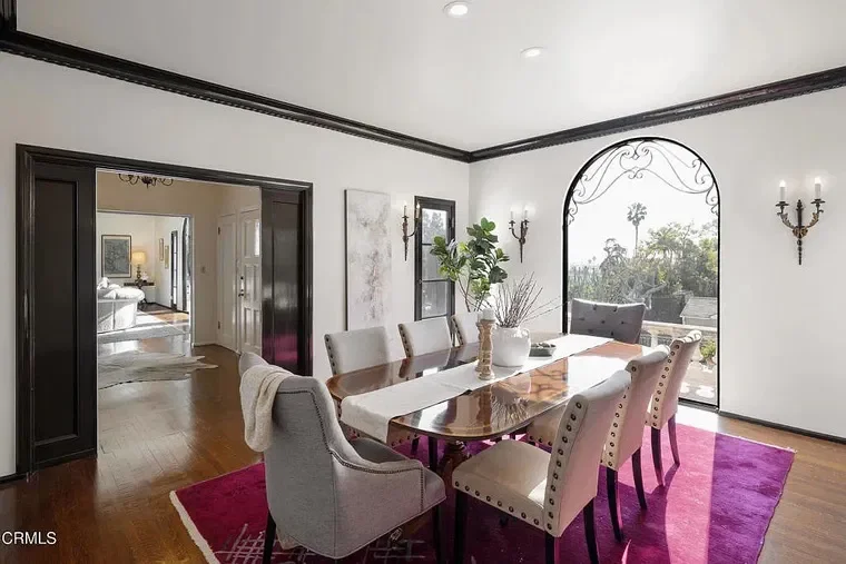 A dining room with a large wooden table, eight upholstered chairs, a white table runner, a vase of greenery, and a large arched window overlooking trees. The room features white walls, dark trim, and a magenta rug under the table. There are wall scon