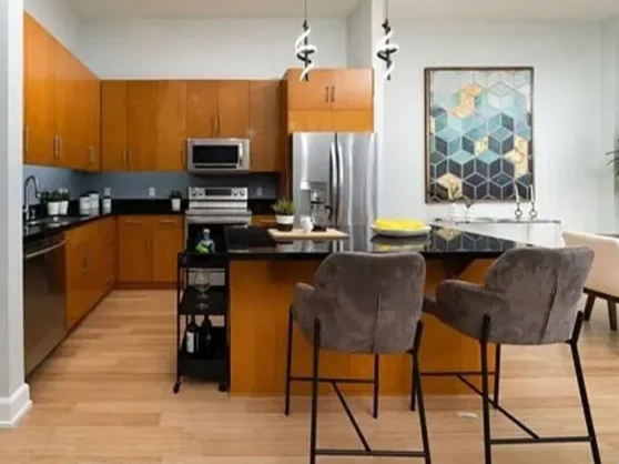 Modern kitchen with wooden cabinets, stainless steel appliances, and a black countertop island. Two gray bar stools are at the island. There is a colorful geometric painting on the wall and pendant lights hanging above the island.