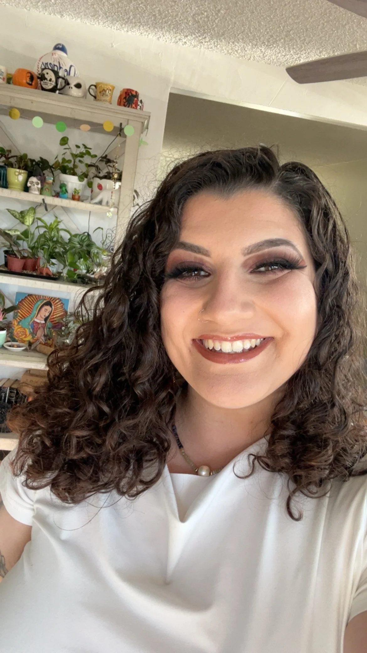 A smiling woman with curly hair, wearing a white top and pearl necklace, taking a selfie in a room decorated with various potted plants, figurines, and artwork on shelves in the background.