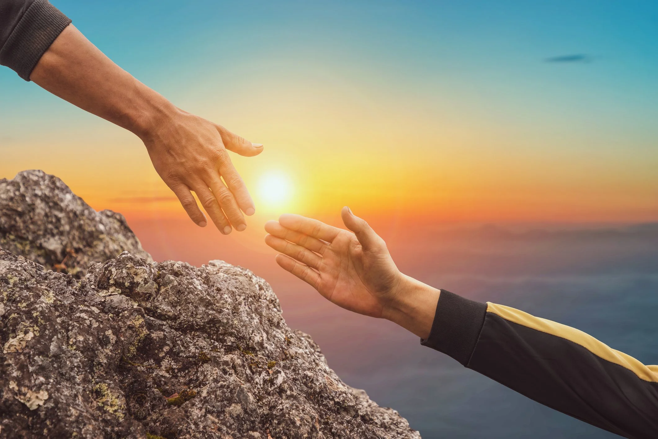 Two hands reaching out to each other over rocks at sunset, one hand extended from a higher position and the other from a lower position.
