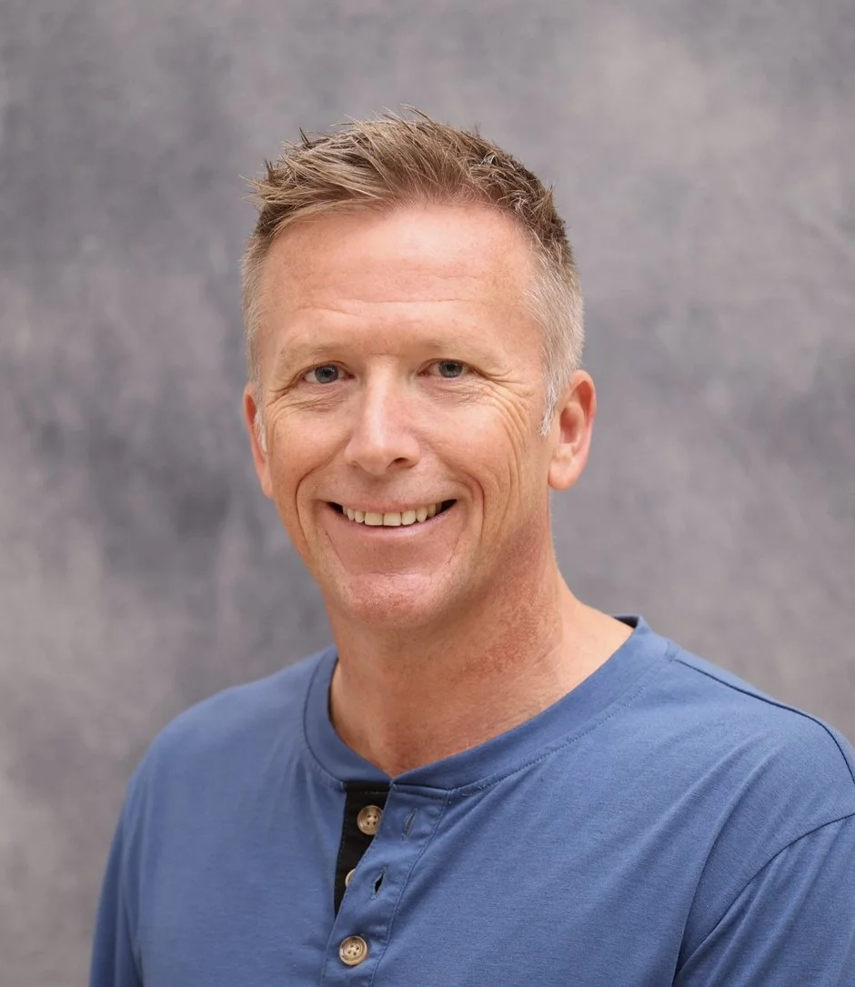 A smiling man with short, light brown hair, wearing a blue shirt, standing against a gray background.