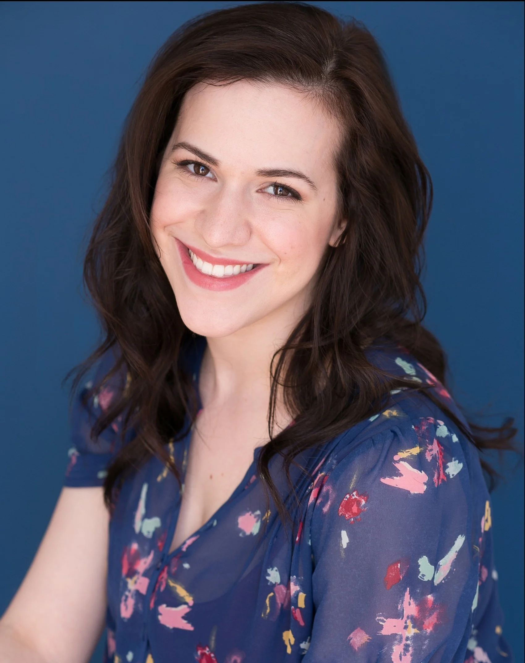 Close-up of a young woman with long, wavy brunette hair smiling against a solid dark blue background. She is wearing a dark blue top with colorful abstract patterns.