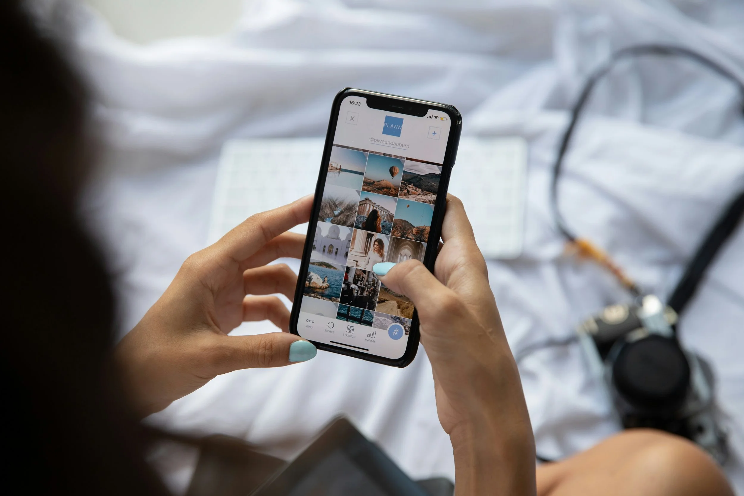 A person holding a smartphone displaying a photo gallery with landscape and travel images, with a camera resting on a white bed in the background.