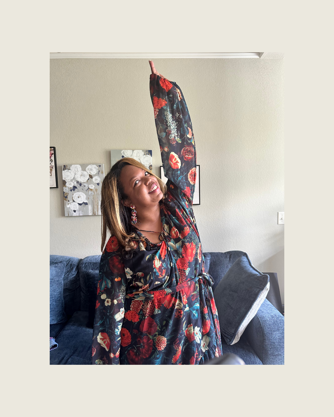A woman wearing a floral dress with red, white, and blue flowers is smiling and stretching her arm upwards in a living room with framed artwork on the wall and a dark gray sofa.