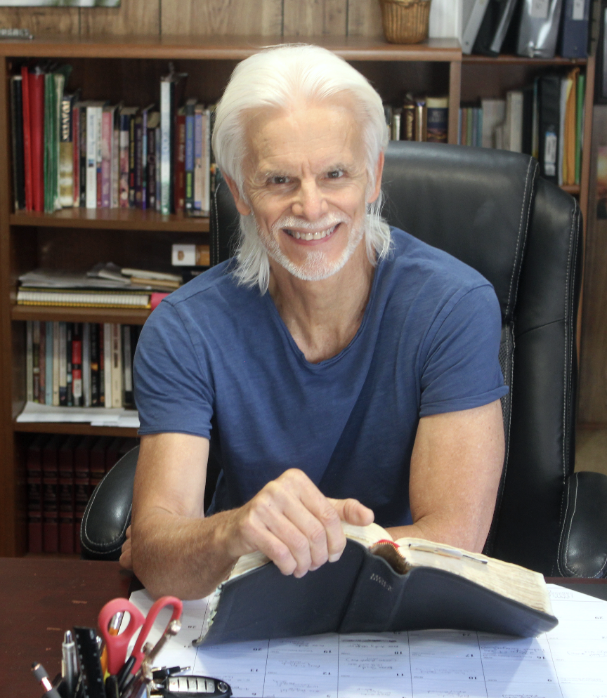An elderly man with white hair and beard, wearing a blue t-shirt, sitting at a desk with a large open book, smiling, in a room with bookshelves in the background.