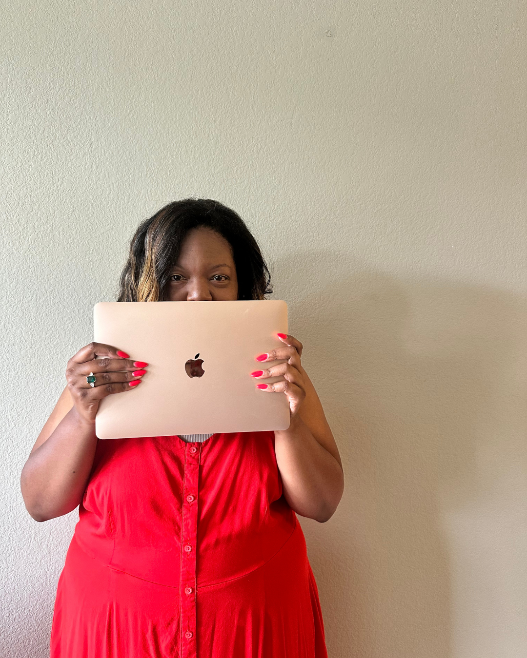 A woman with dark hair, wearing a red dress, holding a rose gold MacBook in front of her face, standing against a plain beige wall.