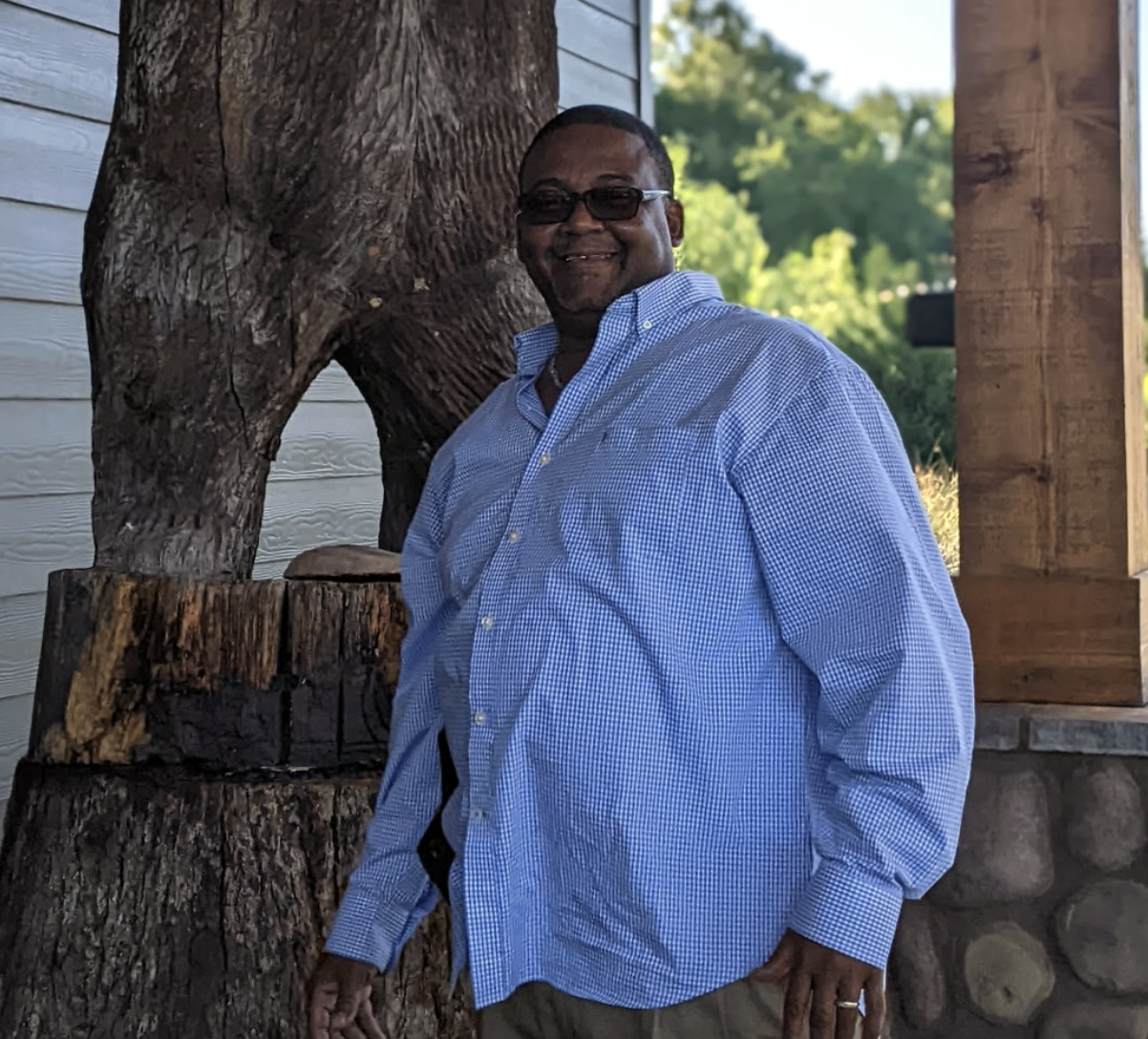 A man with dark skin wearing sunglasses, a blue checkered shirt, and khaki pants standing outdoors next to a large tree and a wooden post.