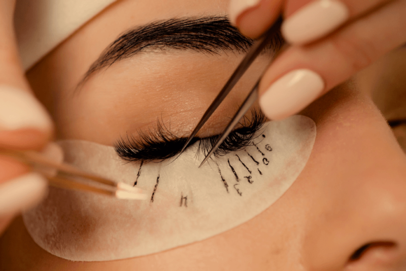 Close-up of a person having eyelash extensions applied using tweezers, with a protective under-eye pad labeled 'L' and 'R' beneath their eye.