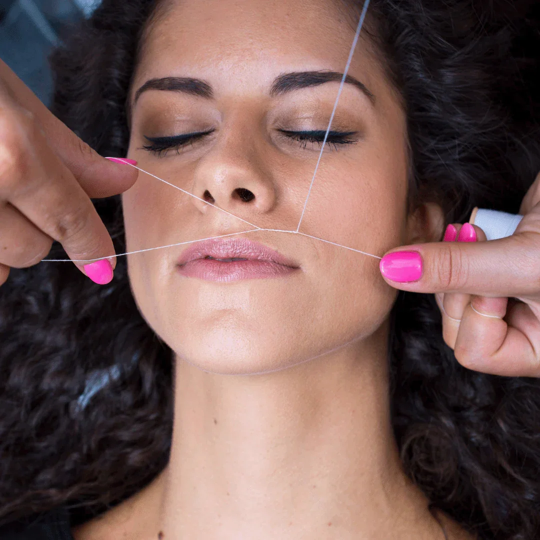 A woman with curly dark hair getting a facial treatment with thin threads placed on her face for contouring, with her eyes closed and pink nail polish.