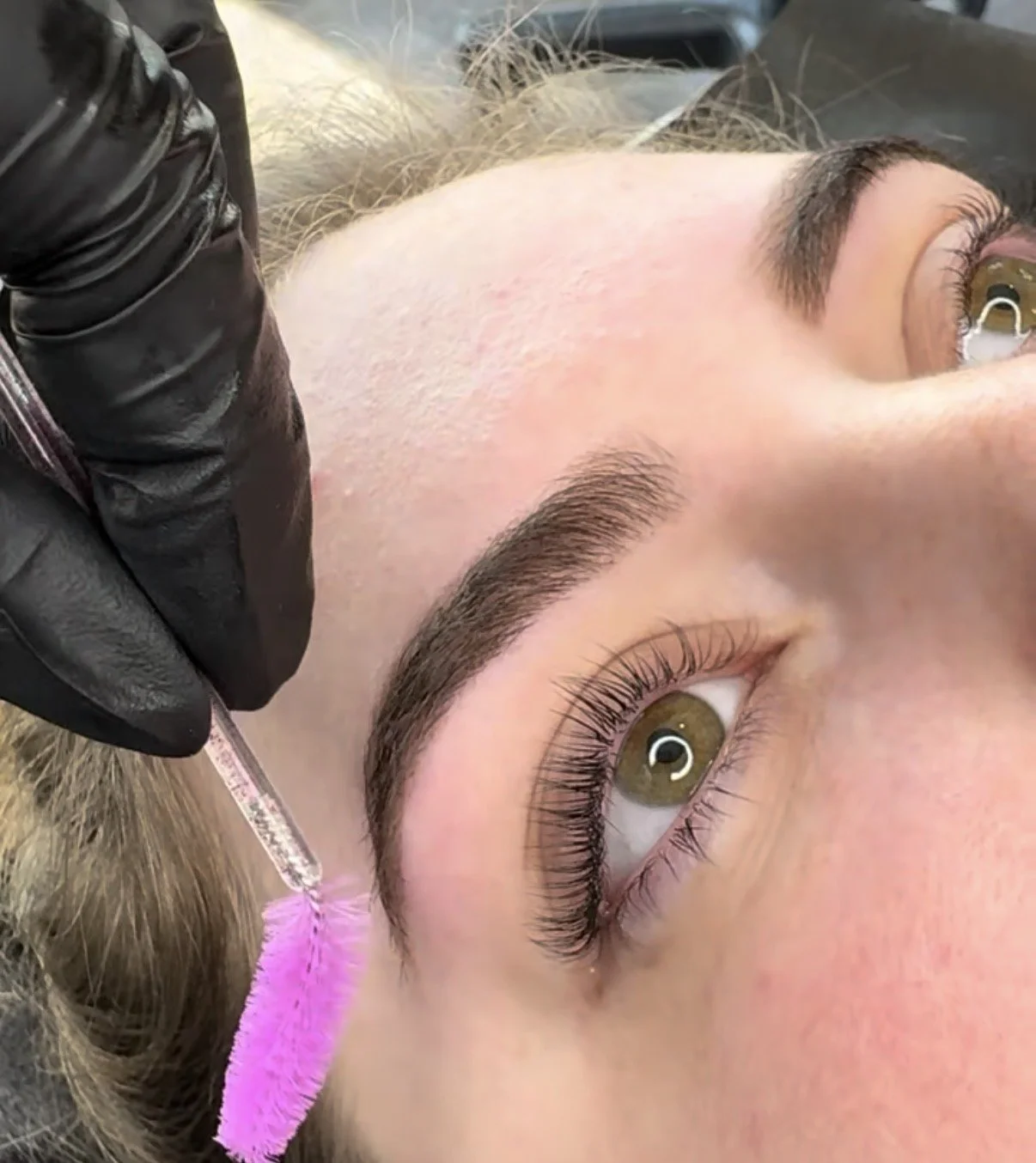 A woman receiving a lash lift treatment, lying down with her eyes open, while a professional in black gloves applies a chemical solution using a small brush on her upper eyelashes.
