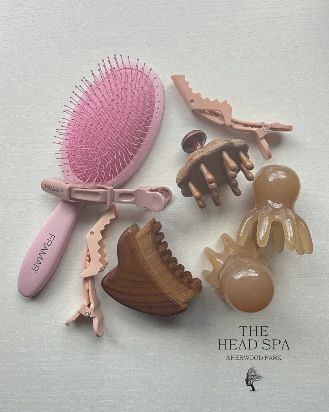A collection of hair accessories and hair clips including a pink scalp massager, a wooden hair clip, and a beige octopus-shaped hair clip, arranged on a white surface with an overlay text "The Head Spa Sherwood Park."