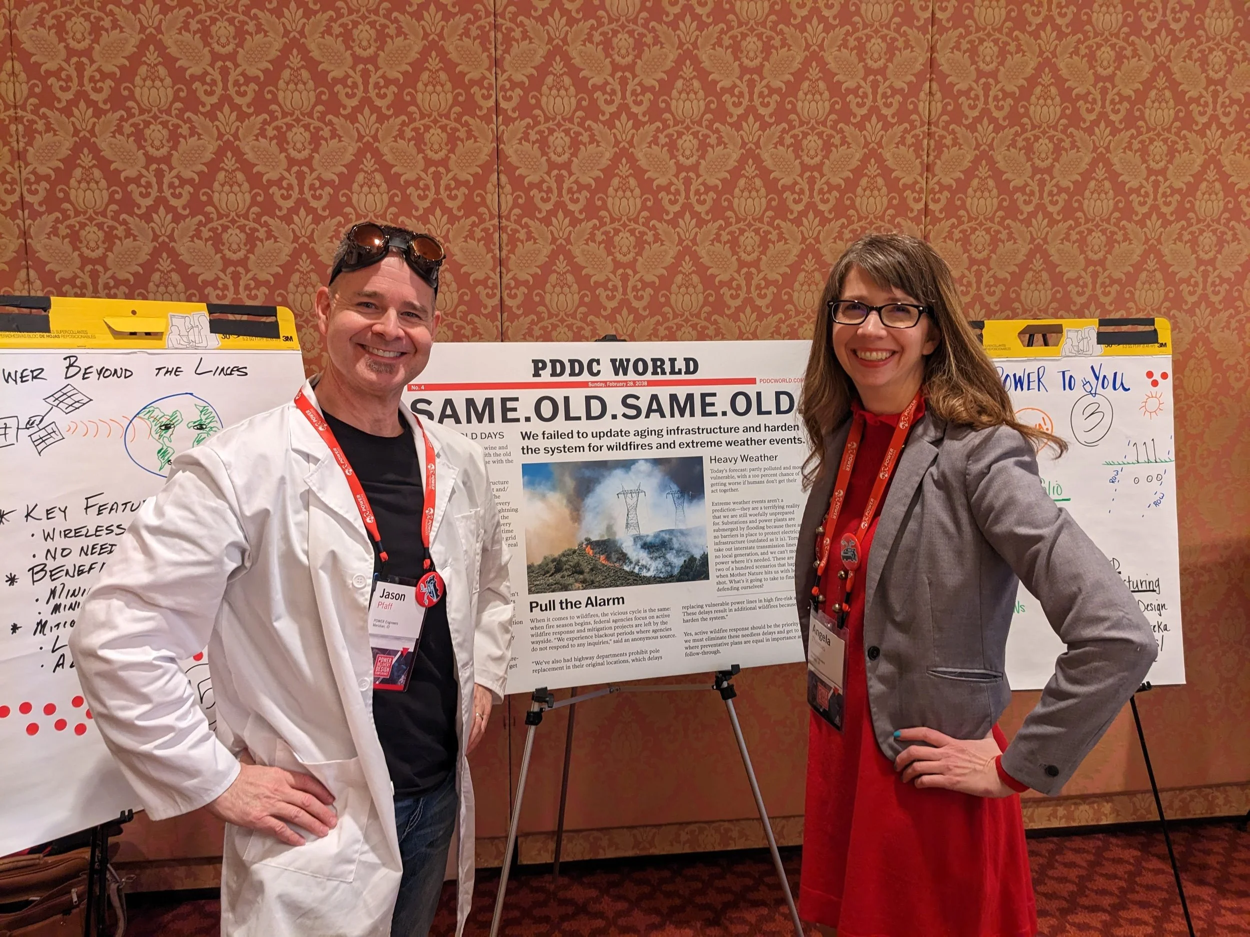 Two smiling professionals, a man and a woman, standing in front of a presentation board with a red ornate patterned wallpaper in the background. The man is wearing a white lab coat and sunglasses on his head, while the woman is wearing glasses and a grey blazer. Both are wearing red conference lanyards.