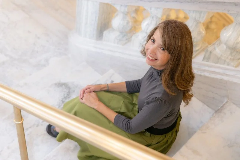 A woman with long reddish-brown hair, wearing a gray top and green skirt, sitting on white marble stairs with a gold-colored railing, smiling up at the camera.