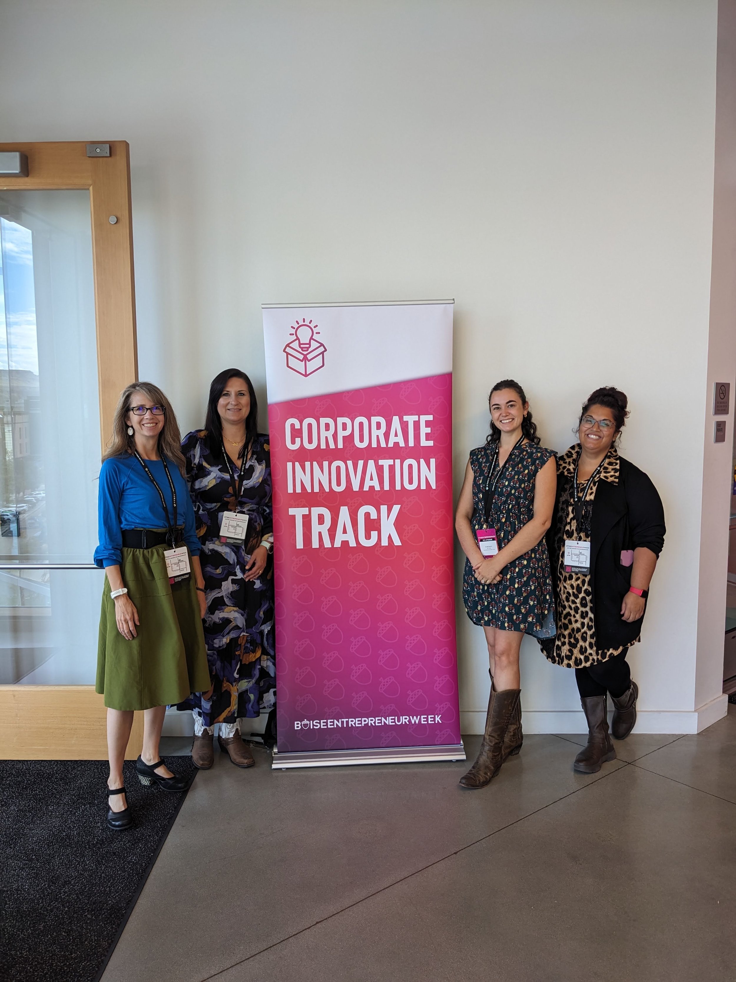 Four women standing together near a pink banner that reads 'Corporate Innovation Track' and '#BoiseEntrepreneurWeek' inside a modern building with a glass door and a window on the left.
