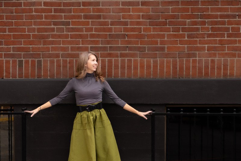 A woman with shoulder-length brown hair, wearing a gray turtleneck and a bright green skirt, standing against a brick wall with a black railing.