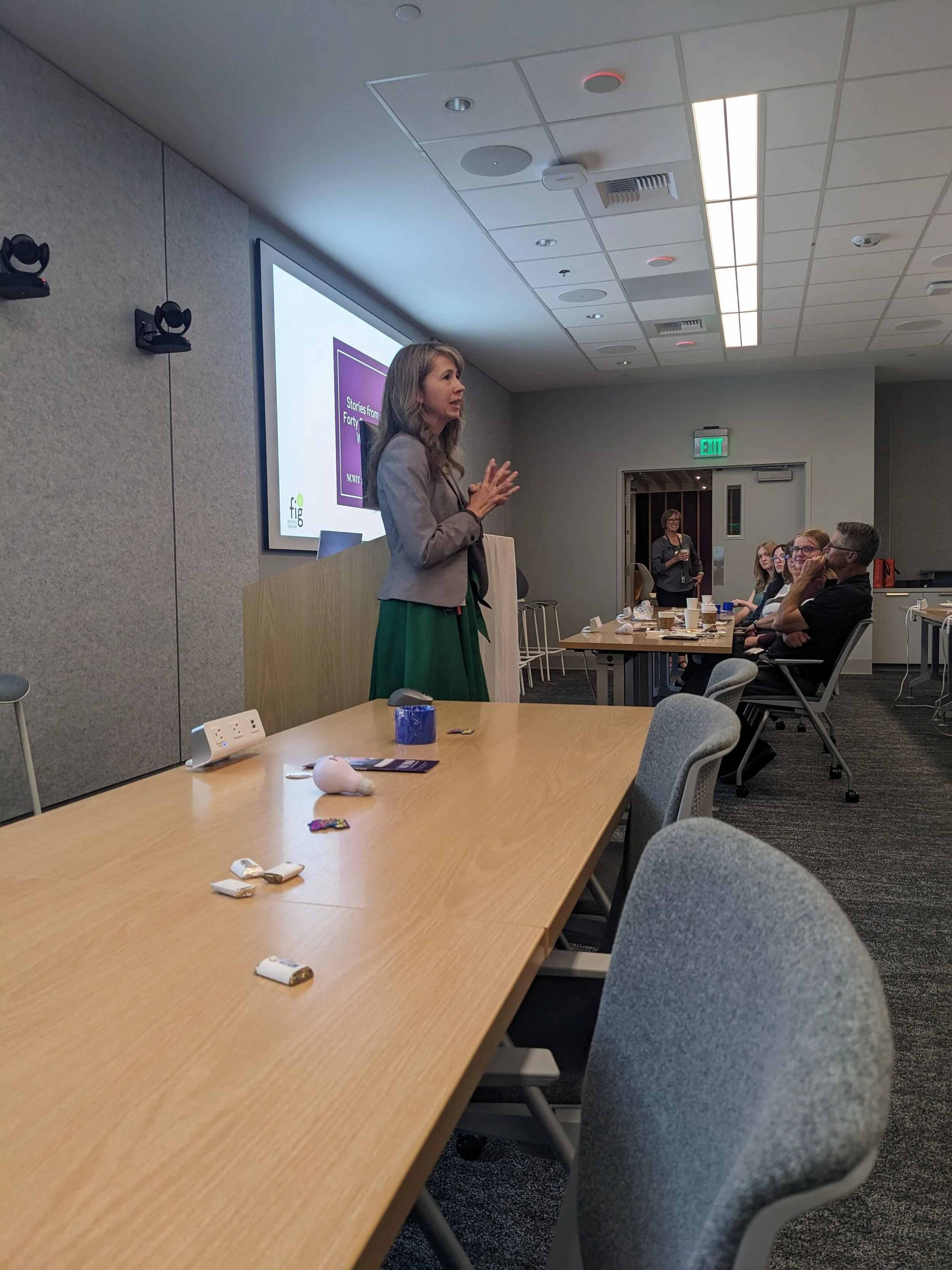 A woman giving a presentation in a conference room, with a group of people seated at tables, some taking notes, while a large screen displays a slide with the text 'Stories from Fortune'.