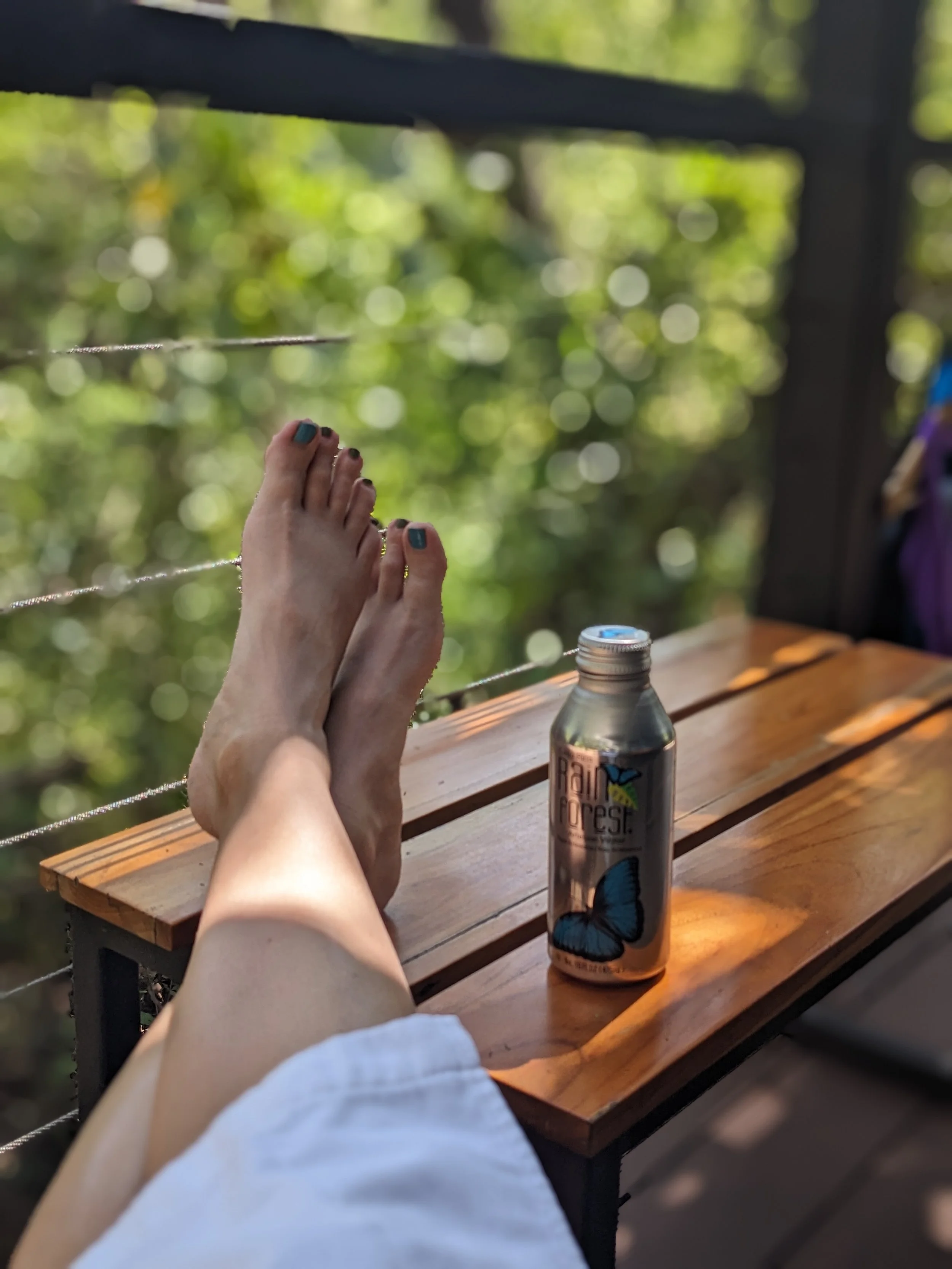 Person with painted toenails relaxing on a wooden outdoor table, with a can of butterfly rain forest drink nearby, surrounded by a garden or forest background.