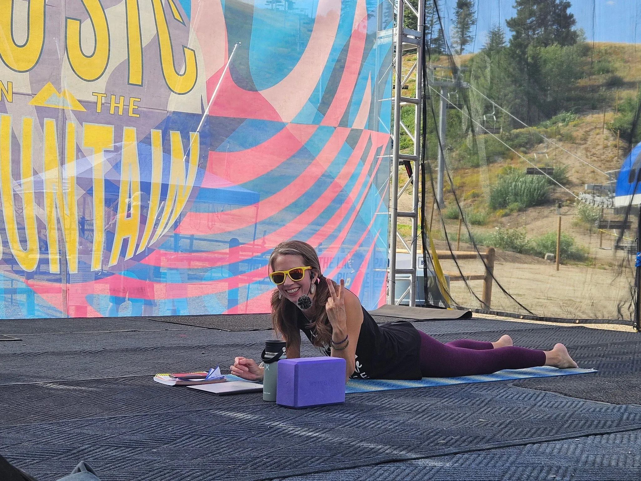 A woman with long hair wearing sunglasses and a black tank top is lying on her stomach on a trampoline outside. She is smiling and making a peace sign with her right hand. Behind her, there is a large, colorful, geometric banner with the words "The" and some other partial text visible. There are some books, a purple box, and a water bottle on the trampoline next to her. The background shows a hilly area with trees and some equipment.