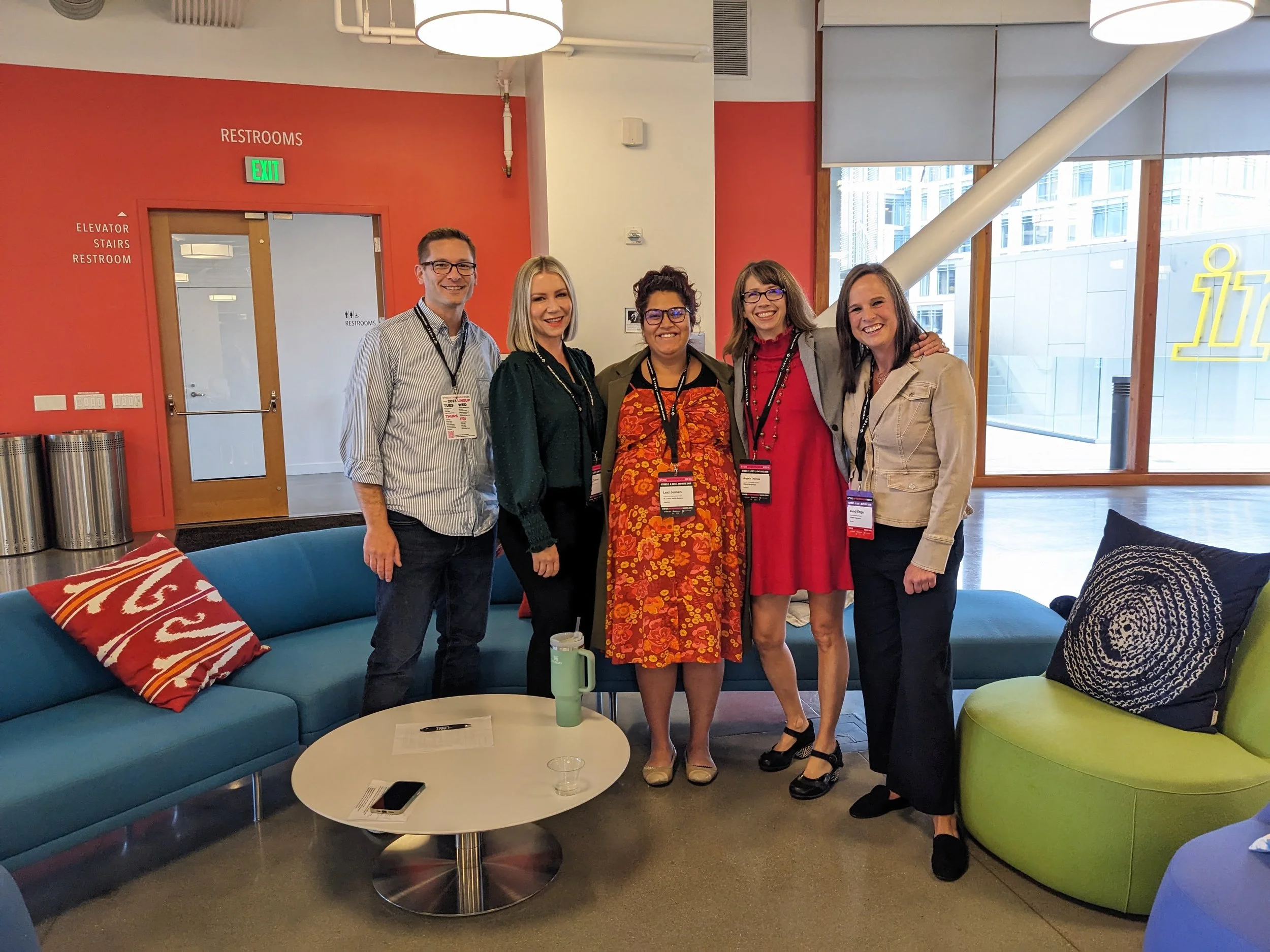 Five women and one man standing together smiling in a modern indoor setting with colorful furniture, white circular light fixtures, and large windows showing city buildings outside.